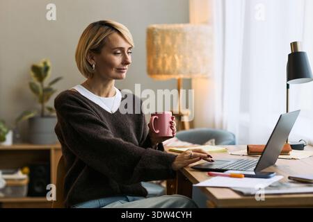 Caucasian middle aged woman sitting at desk using laptop and holding coffee mug, working on femtech health project in home office environment with documents and stationery visible Stock Photo