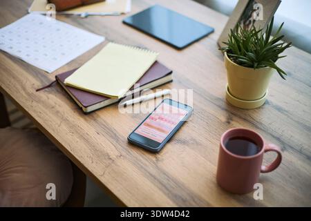 Smartphone displaying femtech health tracking app lying on wooden desk surrounded by notepad, calendar, pen, coffee mug, digital tablet, potted plant, workspace setting Stock Photo