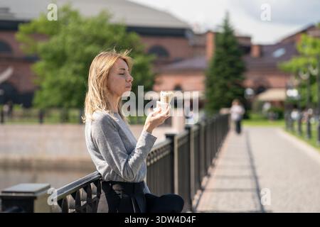 Delightful business woman enjoying taste of ice cream snack while walking outdoors on summer day. Stock Photo