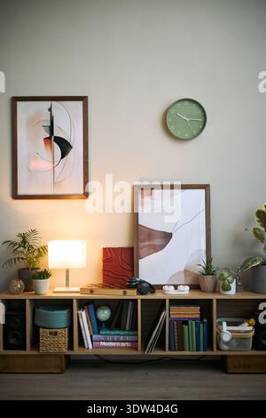 Modern living room shelf displaying books, potted plants, headphones, decorative objects, abstract paintings on wall, green clock above, no people visible, copy space on wall Stock Photo