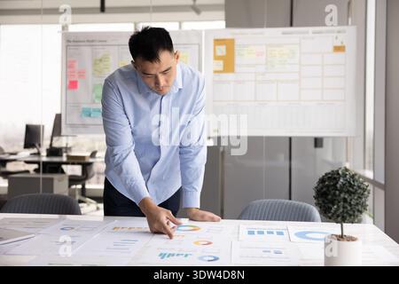Thoughtful young businessman standing at table studying printed finance reports Stock Photo