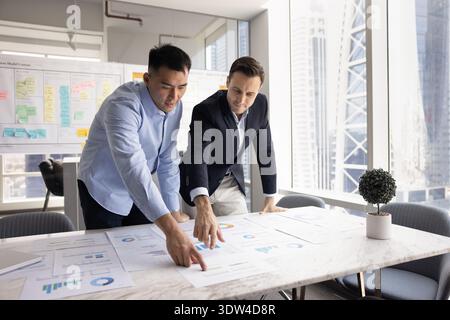 Young businessman standing at table with printed statistics following mentor Stock Photo