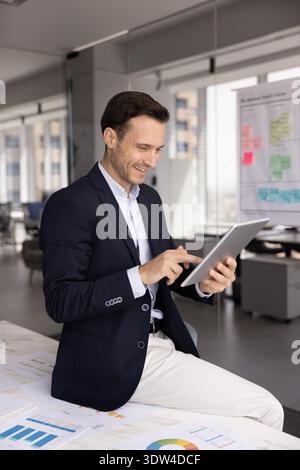 Smiling confident millennial businessman sitting on table using tablet device Stock Photo