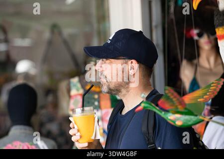BANGKOK, THAILAND-JAN 25, 2026: Man backpack holding plastic cup of orange juice in hand, natural candid motion, side profile,  Chatuchak Market. Stock Photo