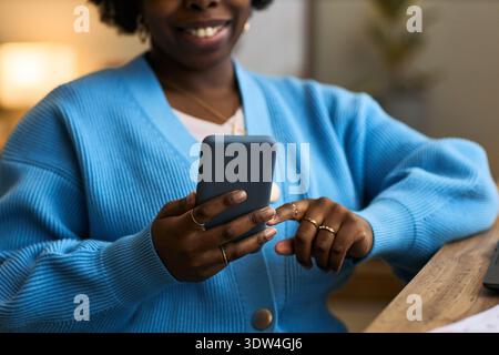 Young adult Black woman using smartphone while sitting at desk, smiling and interacting with mobile health app, showcasing femtech technology in modern healthcare context Stock Photo