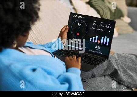 Black woman sitting on sofa using laptop displaying health monitoring dashboard with live vitals, heart rate, sleep analysis and wellness data, representing femtech application Stock Photo