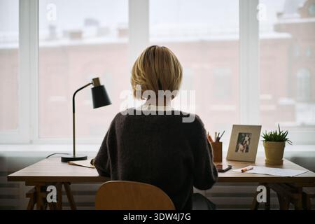 Unrecognizable woman sitting at desk using digital tablet in modern workspace with framed photo and potted plant, engaging in health related activity or working near window Stock Photo