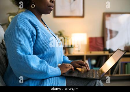 Young adult Black woman sitting indoors using laptop computer for femtech health application, hands typing on keyboard, partial side view showing focused engagement with technology Stock Photo