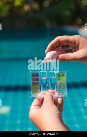 Hand holding pool water test kit measuring pH and chlorine levels in clear blue swimming pool, routine pool maintenance and water quality control Stock Photo