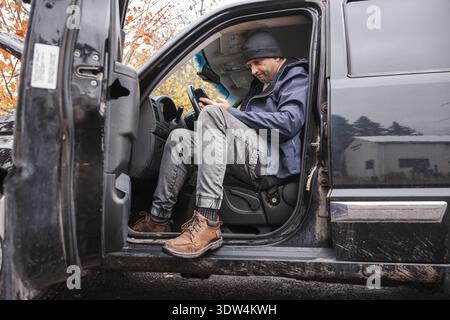 Man sitting in pickup truck and using smartphone during outdoor workday Stock Photo