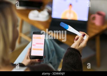 Caucasian woman holding digital ovulation test and smartphone displaying menstrual cycle tracking app, sitting indoors with laptop in background, focusing on femtech health monitoring Stock Photo