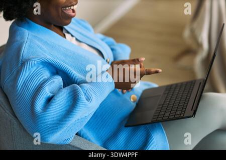 Young adult Black woman sitting and gesturing while using laptop, engaging in remote health consultation or telemedicine session related to femtech, face partially visible Stock Photo