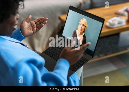 Black woman young adult gesturing while having online video consultation with Caucasian middle aged woman doctor on laptop screen, discussing femtech health concerns in home setting Stock Photo