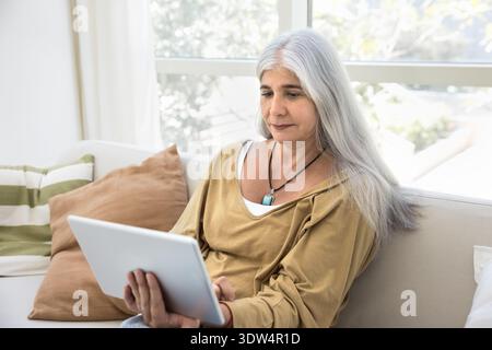 Pretty aged Hispanic woman sitting on couch with digital tablet Stock Photo