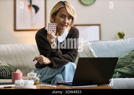 Caucasian woman sitting on sofa holding blister pack of pills while video chatting on laptop, discussing femtech health solutions in home setting, focusing on telemedicine Stock Photo