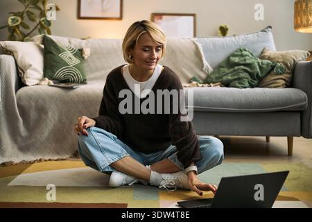 Caucasian young adult woman sitting cross legged on floor using laptop in living room engaging with femtech health application smiling and focusing on screen Stock Photo