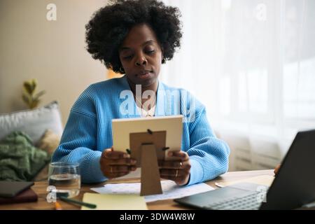 Young adult Black woman sitting at desk holding framed photo and looking at it with nostalgic, thoughtful expression while working with laptop and documents Stock Photo