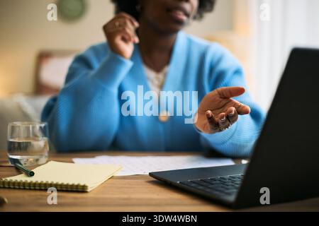 Young adult Black woman gesturing while sitting at desk using laptop for remote health consultation, glass of water and notepad on table, focusing on femtech and wellness Stock Photo