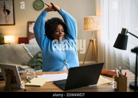 Black woman stretching arms while sitting at desk with laptop and documents, working remotely in home office, promoting femtech health and wellness lifestyle Stock Photo
