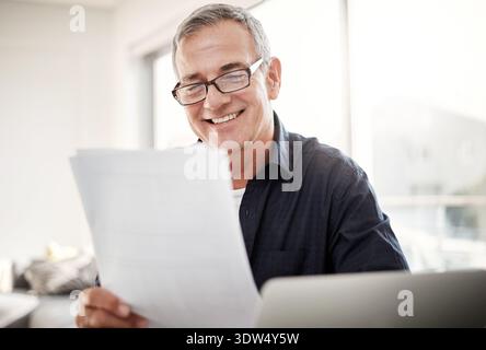 Laptop, document or mature man in home for remote work, invoice review or compare client data. Happy, freelance accountant or tech with papers to Stock Photo
