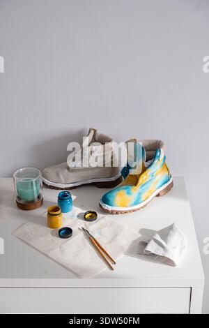 Two boots, one plain and one custom painted with blue and yellow colors, on white table with paints and brushes. Shoe customization. Stock Photo