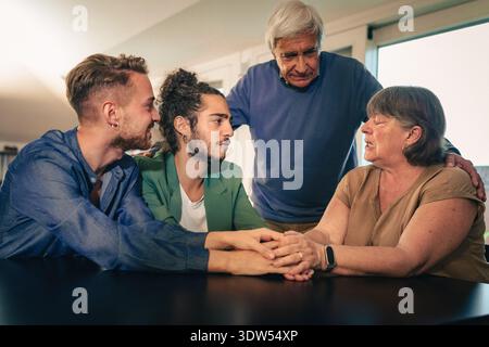 A diverse multi-generational family, including a young gay couple and their elderly parents, sits together at a table indoors, holding hands and shari Stock Photo