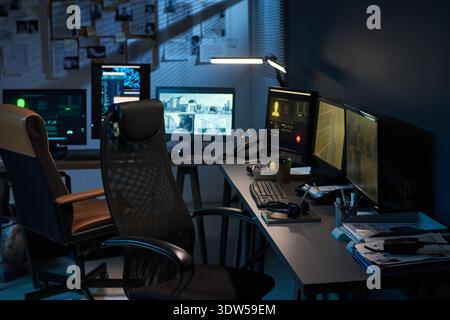 Empty office workspace showing multiple computer monitors displaying surveillance footage and data analysis screens, ergonomic chair positioned in front of desk with electronic devices and documents Stock Photo