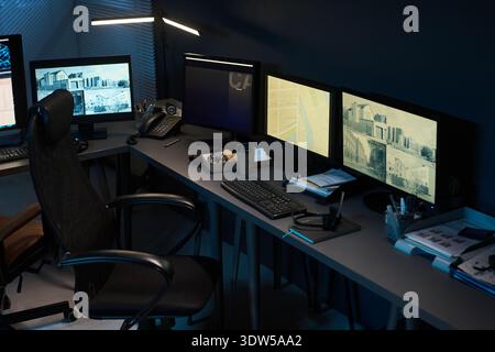 Empty modern office workspace featuring multiple computer monitors displaying surveillance footage, keyboard, mouse, telephone, coffee cup, and office supplies on desk Stock Photo