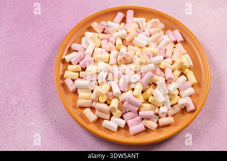 Mini colorful marshmallows piled on an orange ceramic plate, creating a playful, pastel arrangement of pink, yellow and white sweets against a texture Stock Photo