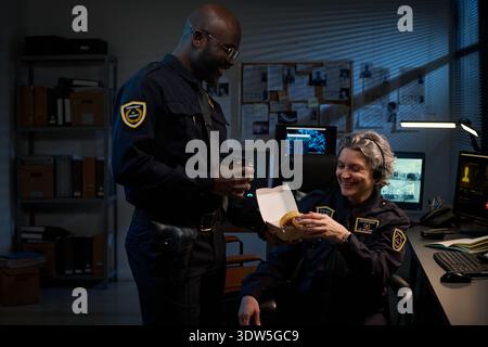 Young Black man standing and holding coffee cup, smiling at seated middle aged Caucasian woman offering donut in office with computer monitors and files visible Stock Photo