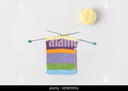 Striped piece of knitted fabric for future clothing with knitting needles and yellow ball of thread on gray background. Homemade needlework and knitting of warm, clothes and toys. Concept creativity Stock Photo