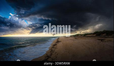 View of ominous dark storm clouds roll in over the sandy beach and turbulent sea, contrasting with the golden light, Taranto, Apulia, Italy. Stock Photo