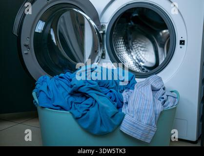 Basket filled with dirty blue and striped clothes placed in front of an open washing machine. Domestic interior scene showing preparation for laundry. Stock Photo