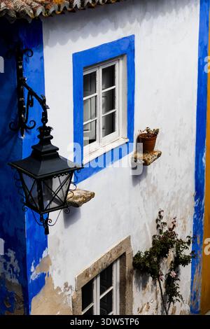 View of a quaint building facade with brilliant white walls framed by striking blue accents, a charming window, and an ornate lamp, Óbidos, Leiria Dis Stock Photo