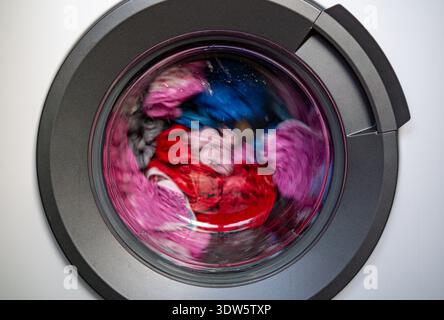 View through the round glass door of a front loading washing machine with colorful clothes spinning during the wash cycle. Stock Photo