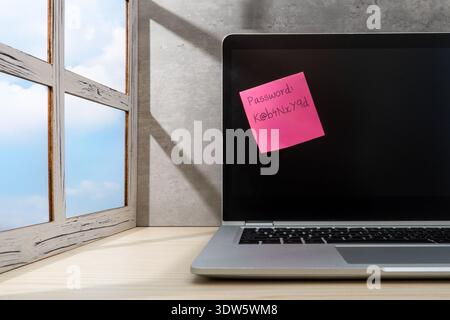 A note with a complex password written on it is attached to a laptop screen. Cybersecurity, data privacy risks, password management concept. Stock Photo