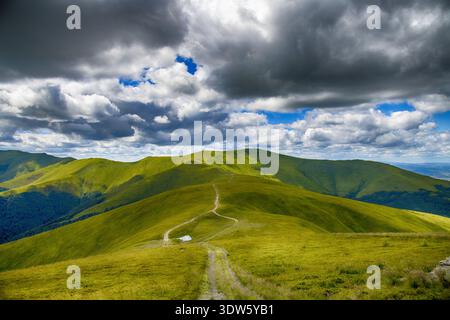 View of a winding dirt trail cutting through vibrant green hills under a dramatic sky with dark clouds and patches of blue, Pylypets', Zakarpattia Obl Stock Photo