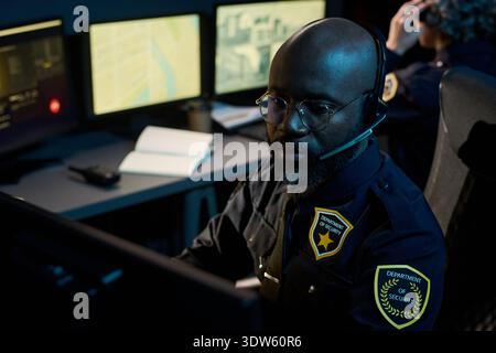 Black man wearing headset working as security officer in control room monitoring multiple computer screens with maps and data, focused on surveillance operations Stock Photo