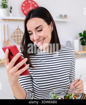 Happy woman eating fresh salad and browsing social media on phone in kitchen Stock Photo
