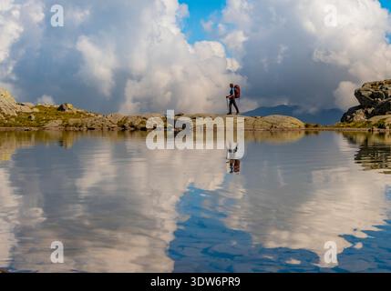 A hiker explores a serene alpine landscape in Swizz, surrounded by towering clouds and a calm lake. The reflection creates a picturesque scene of Stock Photo