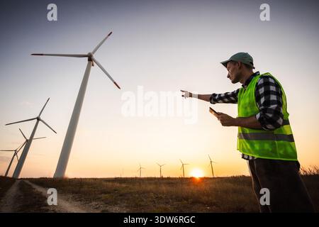 Engineer in safety vest pointing towards wind turbines at sunset, concept of renewable energy development and sustainable power control. Stock Photo