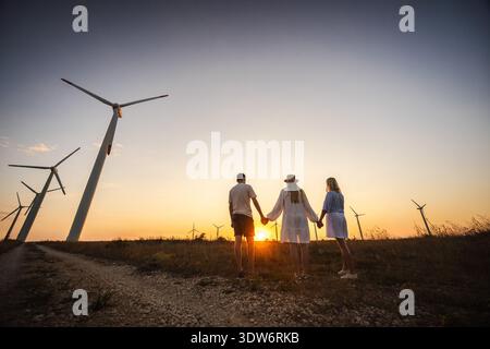 Family holding hands at sunset near wind turbines, symbolizing unity, sustainability, and future generations. Stock Photo