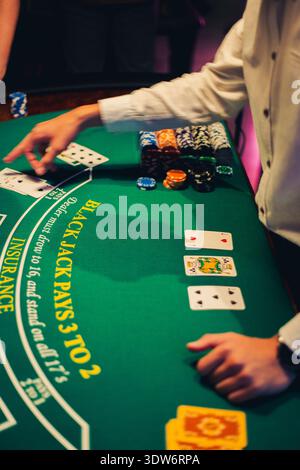 A close-up view of a blackjack table with playing cards, colorful stacks of casino chips and a dealer’s hand indicating gameplay. Stock Photo