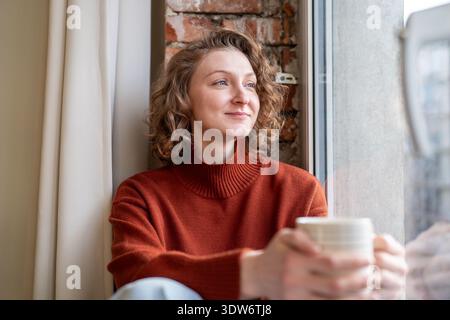 Thoughtful serene young woman enjoys moment of mindfulness with cup of coffee take break near window Stock Photo