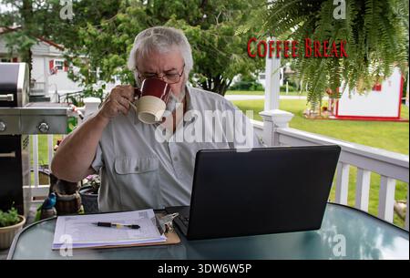 A man sits at a table on a porch, holding a cup of coffee. He uses a laptop to work. A grill and a bright yard are visible around him as he takes a break in the daytime. Stock Photo