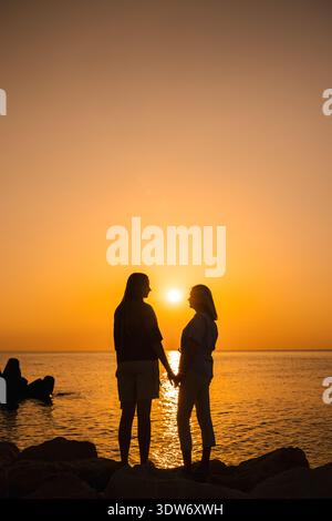 Two women standing on rocks by the sea at sunrise, holding hands and facing each other, symbolizing love, family, emotional wellbeing Stock Photo