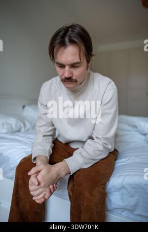 Depressed frustrated man sitting on unmade bed, hands nervously clasped in tension deep in thoughts. Stock Photo