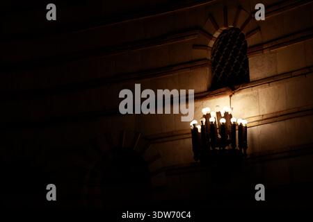 A vintage chandelier with multiple illuminated bulbs casts a warm glow on the textured stone walls and arched window of a dimly lit room Stock Photo