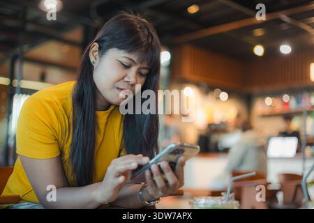 Young Asian woman in yellow shirt using smartphone in a cafe. Concept of social media marketing, mobile application usage, digital communication, onli Stock Photo
