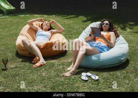 Two young women relaxing on bean bag chairs in a green garden. One is sunbathing while the other uses a digital tablet on a sunny summer day. Stock Photo